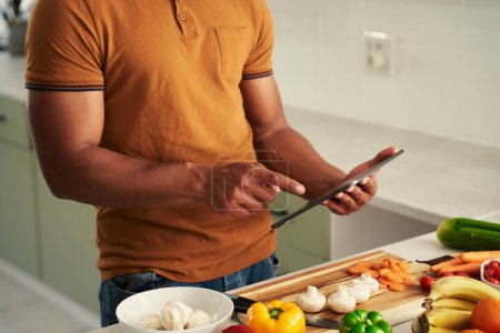 Foto de Primer plano del joven birracial que usa ropa casual usando tableta digital junto a la comida en la cocina - Imagen libre de derechos