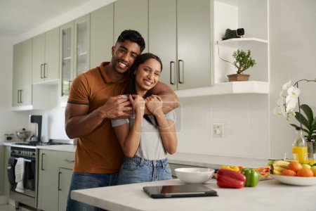 Foto de Feliz joven pareja biracial sonriendo y abrazando junto a la comida y la tableta digital en la cocina - Imagen libre de derechos