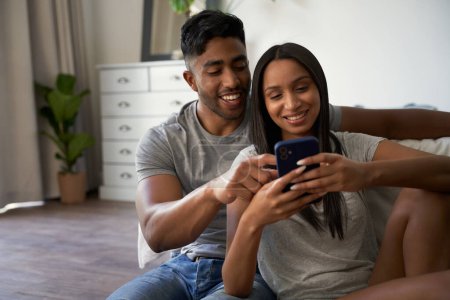 Téléchargez les photos : Heureux jeune couple biracial portant des vêtements décontractés en utilisant le téléphone portable dans la chambre à coucher à la maison - en image libre de droit