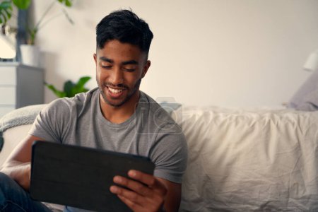 Téléchargez les photos : Heureux jeune homme biracial portant des vêtements décontractés en utilisant une tablette numérique dans la chambre à coucher à la maison - en image libre de droit