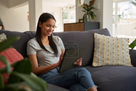 Foto de Feliz joven biracial mujer en ropa casual utilizando tableta digital en el sofá en la sala de estar - Imagen libre de derechos
