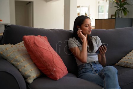 Téléchargez les photos : Jeune femme biraciale portant des vêtements décontractés en utilisant un téléphone portable et des écouteurs sans fil sur le canapé à la maison - en image libre de droit