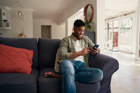 Photo for Happy young biracial man wearing casual clothing sitting on sofa using mobile phone at home - Royalty Free Image