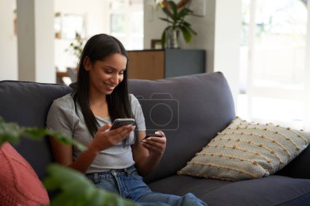 Foto de Mujer biracial joven con ropa casual sonriendo mientras usa el teléfono móvil para la banca en el sofá en casa - Imagen libre de derechos