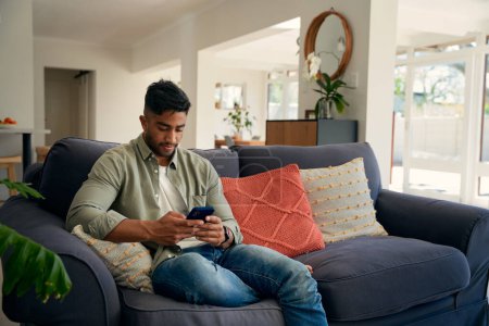 Photo for Young biracial man wearing casual clothing sitting on sofa typing on mobile phone at home - Royalty Free Image