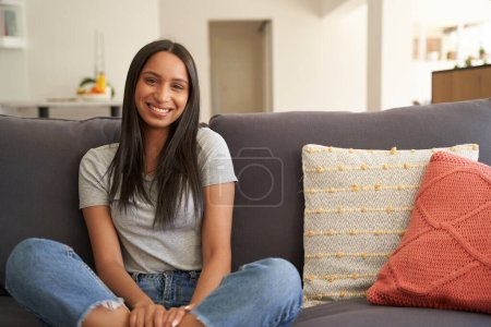 Téléchargez les photos : Joyeux jeune femme biraciale portant des vêtements décontractés souriant tout en étant assis sur le canapé dans le salon à la maison - en image libre de droit