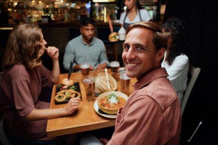 Foto de Feliz joven grupo multirracial de amigos que usan ropa casual sonriendo mientras reciben la cena en el bar - Imagen libre de derechos