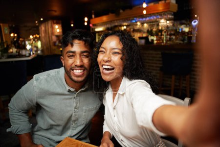 Téléchargez les photos : Jeune couple multiracial portant des vêtements décontractés souriant tout en prenant selfie avec téléphone portable au bar - en image libre de droit