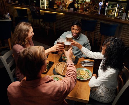 Foto de Feliz joven grupo multirracial de amigos con ropa casual haciendo tostadas con cervezas durante la cena en el bar - Imagen libre de derechos