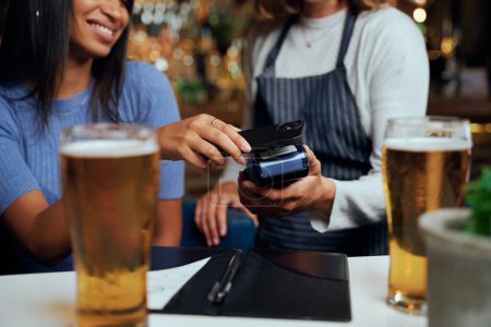 Young woman wearing casual clothing paying for dinner with wireless technology next to waitress
