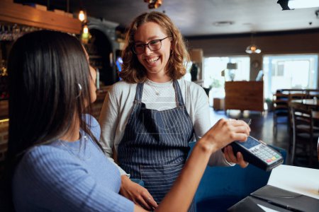 Foto de Mujer joven y feliz vistiendo ropa casual pagando la cena con la máquina de tarjetas por la camarera - Imagen libre de derechos