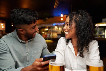 Téléchargez les photos : Jeune couple multiracial portant des vêtements décontractés souriant face à face avec téléphone portable au bar - en image libre de droit