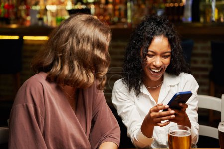 Téléchargez les photos : Heureux jeunes amis multiraciaux portant des vêtements décontractés souriant tout en utilisant le téléphone mobile au restaurant - en image libre de droit