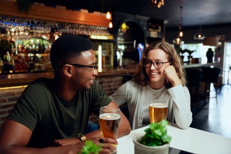 Foto de Felices jóvenes amigos multirraciales usando ropa casual sonriendo cara a cara con cervezas en el bar - Imagen libre de derechos