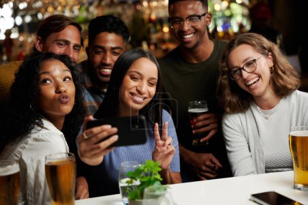Foto de Feliz joven grupo multirracial de amigos que usan ropa casual tomando selfie con teléfono móvil en el bar - Imagen libre de derechos