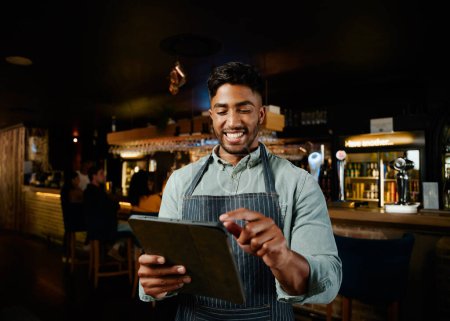 Foto de Feliz joven biracial hombre con delantal apuntando a la tableta digital mientras trabaja como camarero en el bar - Imagen libre de derechos