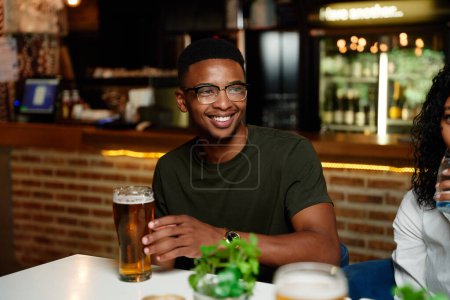 Foto de Felices jóvenes amigos multirraciales usando ropa casual sonriendo y bebiendo cerveza en el bar - Imagen libre de derechos