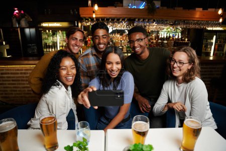 Téléchargez les photos : Heureux jeune groupe multiracial d'amis portant des vêtements décontractés prendre selfie avec téléphone portable au restaurant - en image libre de droit