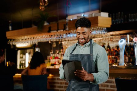 Téléchargez les photos : Heureux jeune homme biracial portant tablier en utilisant une tablette numérique tout en travaillant comme serveur dans le restaurant - en image libre de droit