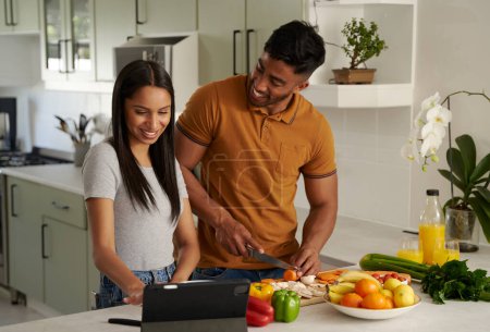 Foto de Feliz joven pareja biracial con ropa casual preparando comida al lado de la tableta digital en la cocina - Imagen libre de derechos