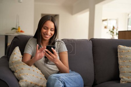 Foto de Mujer biracial joven usando ropa casual usando teléfono móvil mientras está sentada en el sofá en casa - Imagen libre de derechos