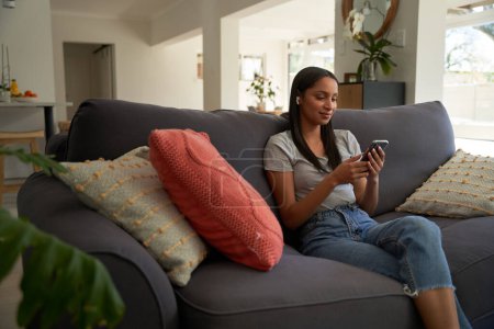 Photo for Young biracial woman wearing casual clothing with mobile phone and wireless earphones on sofa at home - Royalty Free Image
