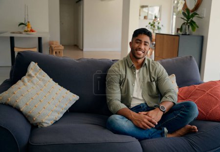Foto de Feliz joven biracial vestido con ropa casual sonriendo mientras está sentado en el sofá en la sala de estar en casa - Imagen libre de derechos