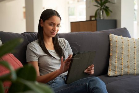 Téléchargez les photos : Satisfait jeune femme biraciale portant des vêtements décontractés en utilisant une tablette numérique sur le canapé dans le salon - en image libre de droit