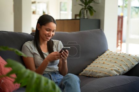 Foto de Mujer biracial joven con ropa casual sonriendo mientras escribe en el teléfono móvil en el sofá en casa - Imagen libre de derechos