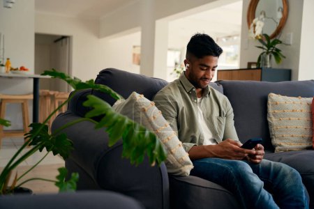 Photo for Young biracial man sitting on sofa using mobile phone and wireless earphones at home - Royalty Free Image