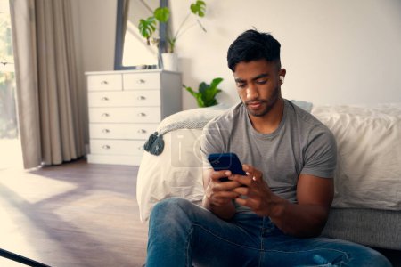 Téléchargez les photos : Jeune homme biracial portant des vêtements décontractés en utilisant un téléphone portable et des écouteurs sans fil dans la chambre à coucher à la maison - en image libre de droit