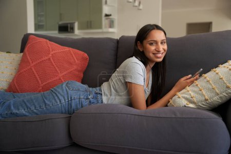 Foto de Feliz joven biracial mujer usando ropa casual acostado en el sofá con el teléfono móvil en casa - Imagen libre de derechos