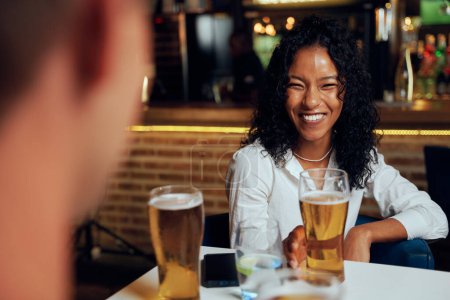Foto de Jóvenes amigos multirraciales que usan ropa casual sonriendo mientras disfrutan de cervezas en el restaurante - Imagen libre de derechos