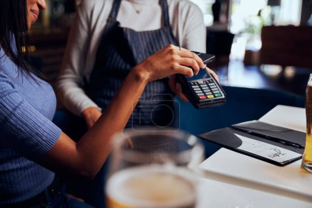 Photo for Young woman wearing casual clothing paying for dinner with card machine next to waitress - Royalty Free Image