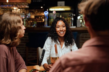 Téléchargez les photos : Heureux jeune groupe multiracial d'amis portant des vêtements décontractés parler pendant le dîner au restaurant - en image libre de droit