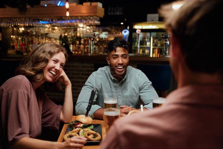 Foto de Joven grupo multirracial de amigos que usan ropa casual riéndose alrededor de la mesa en el restaurante - Imagen libre de derechos