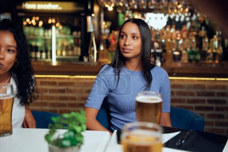 Téléchargez les photos : Jeunes femmes portant des vêtements décontractés regardant loin tout en étant assis à table avec des bières et facture au restaurant - en image libre de droit