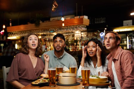 Téléchargez les photos : Choqué jeune groupe multiracial d'amis portant des vêtements décontractés profiter du dîner et des bières au restaurant - en image libre de droit