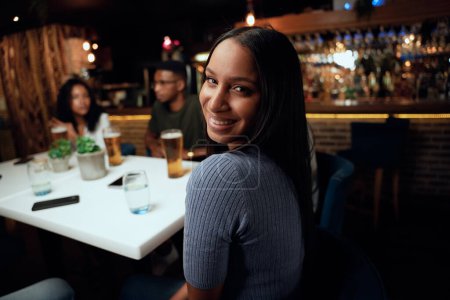 Foto de Feliz joven grupo multirracial de amigos con ropa casual sonriendo y hablando mientras celebran en el bar - Imagen libre de derechos