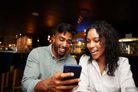 Foto de Joven pareja multirracial que usa ropa casual riéndose y usando un teléfono inteligente en el bar - Imagen libre de derechos