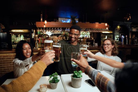 Foto de Feliz joven grupo multirracial de amigos con ropa casual tostadas con bebidas en la mesa - Imagen libre de derechos