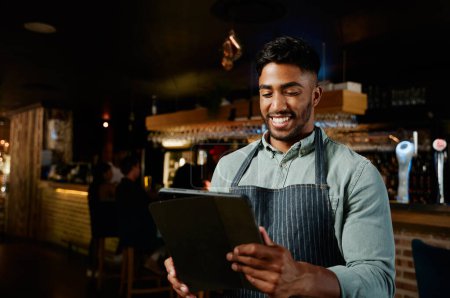 Téléchargez les photos : Jeune homme biracial portant tablier souriant tout en utilisant une tablette numérique tout en travaillant comme serveur dans le restaurant - en image libre de droit