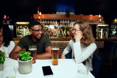 Téléchargez les photos : Heureux jeune groupe multiracial d'amis portant des vêtements décontractés riant avec des boissons à table au bar - en image libre de droit