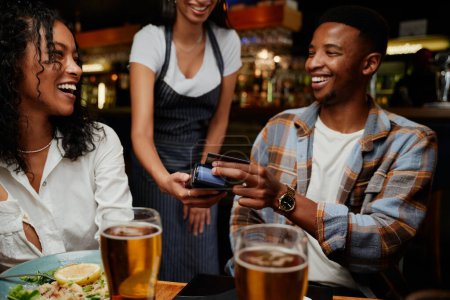 Téléchargez les photos : Heureux amis multiraciaux portant des vêtements décontractés serveuse payante pour le dîner et les boissons au restaurant - en image libre de droit