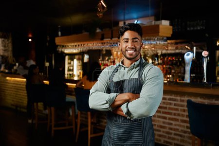 Photo for Young biracial man wearing apron smiling with arms crossed while working as waiter in restaurant - Royalty Free Image
