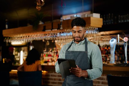 Téléchargez les photos : Jeune homme biracial portant tablier en utilisant une tablette numérique tout en travaillant comme serveur dans le restaurant - en image libre de droit