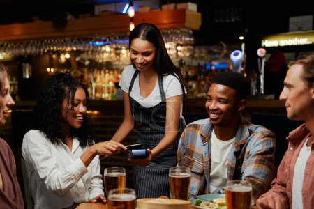 Junge multiethnische Gruppe von Freunden in lässiger Kleidung zahlt für Abendessen und Getränke im Restaurant