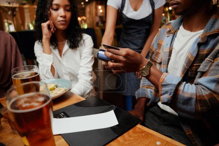 Téléchargez les photos : Gros plan d'amis multiraciaux portant des vêtements décontractés payant la serveuse pour le dîner et les boissons facture au bar - en image libre de droit