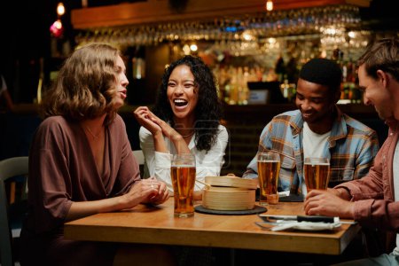 Foto de Feliz joven grupo multirracial de amigos con ropa casual hablando y riendo durante la cena en el bar - Imagen libre de derechos