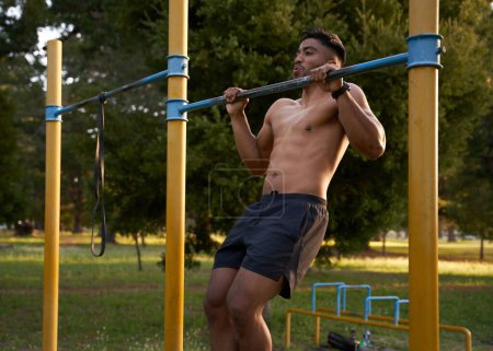 Photo for Shirtless young biracial man doing chin-ups on pull-up bar by trees in the park - Royalty Free Image